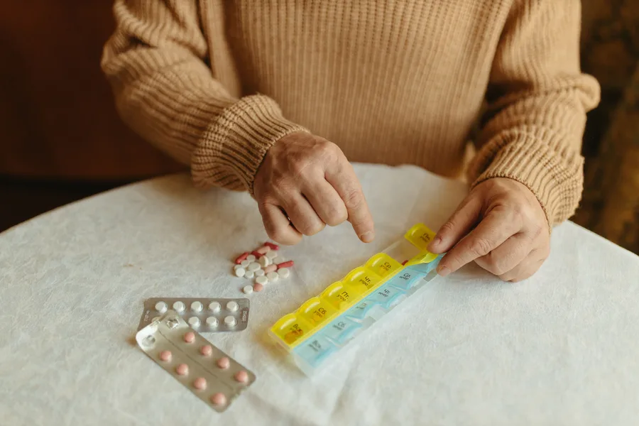 Older person sorting tablets into a weekly pill organiser beside blister packs